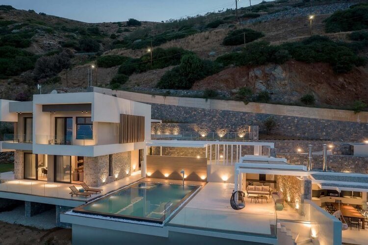 Surrounded by hills, this modern villa is lit up in warm orange with an infinity pool, dining area and sun loungers.