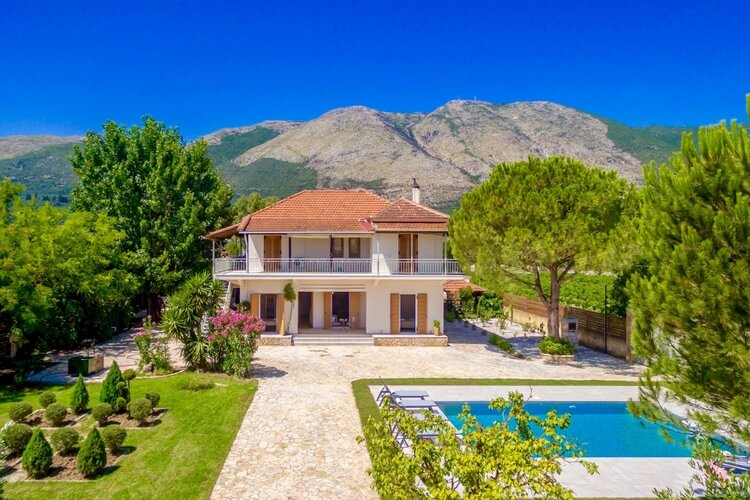 White and orange villa with mountains in the background and a vast garden with a pool in the foreground