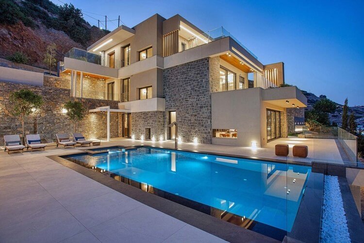 A beautiful white and stone modern villa at the foot of the hill. There is an infinity pool painted at dusk and illuminated in the foreground.